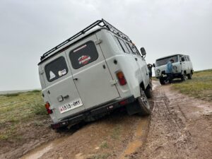 Two vehicles in muddy terrain: one vehicle is deeply stuck in a mud rut with its rear end tilted, and the other is positioned better with a person inspecting or assisting.